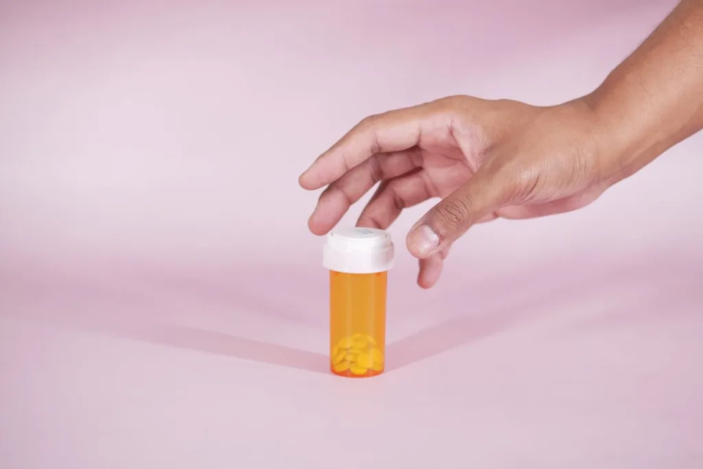 A hand reaches for a prescription bottle with yellow pills on a pink background, symbolizing healthcare.